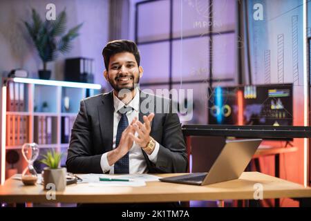 Primo piano ritratto di sicuro indiano esperto finanziario ufficio lavoratore seduto a un tavolo in abito formale lavorare con il computer portatile in ufficio moderno sera, battendo le mani. Foto Stock