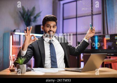Concetto di lavoro a distanza di notte. Attraente fiducioso arabo esperto barbuto uomo d'affari che spiega grafico economico su lavagna di vetro utilizzando il portatile in ufficio serale mostrando segno ok, okay. Foto Stock