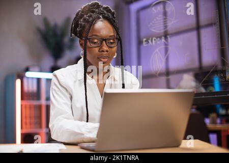 Donna afroamericana in abbigliamento formale seduto alla scrivania e guardando un moderno computer portatile con seria espressione facciale. Concetto di persone e lavoro in ufficio. Foto Stock