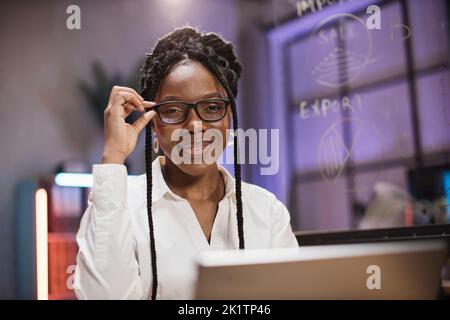 Donna afroamericana in abbigliamento formale seduto alla scrivania e guardando un moderno computer portatile con seria espressione facciale. Concetto di persone e lavoro in ufficio. Foto Stock