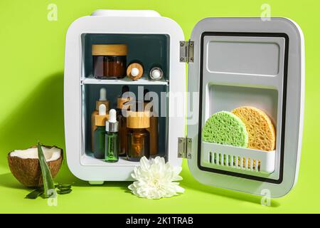 Piccolo frigorifero con cosmetici naturali, cocco, foglie di aloe e fiori su sfondo verde Foto Stock