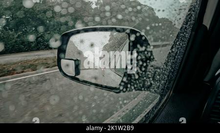 Specchietto retrovisore lato guida con goccioline di pioggia. Guidare l'automobile mentre il tempo piovoso, strada trafficata Foto Stock