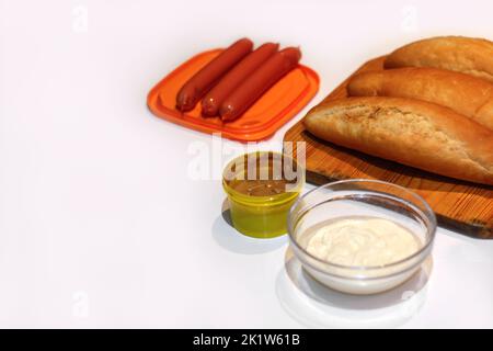 Ingredienti sfocati per preparare hot dog fatti in casa. Salsicce in piatto d'arancia, panini appena sfornati, senape e salsa su fondo bianco. Hotdog con diffe Foto Stock