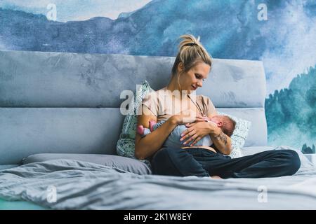 Donna bionda bianca seduta a letto in una camera da letto blu che tiene il bambino tra le braccia allattandolo con espressione affettuosa. Maternità. Scatto orizzontale in interni. Foto di alta qualità Foto Stock