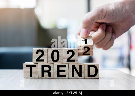 Mano di businessman che tiene cubo di legno con il blocco di ribaltamento 2021 - 2022 PAROLA DI TENDENZA sullo sfondo della tabella. Risoluzione, strategia, soluzione, obiettivo, business Foto Stock