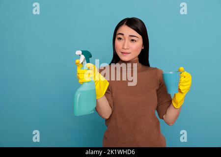 Triste depresso giovane governante asiatico in possesso di uno spray e caffè tazza indossando guanti gialli, pulizia concetto casa, tè di rinfresco pausa tempo di servizio, infelice esaurito e negativo sensazione donna Foto Stock