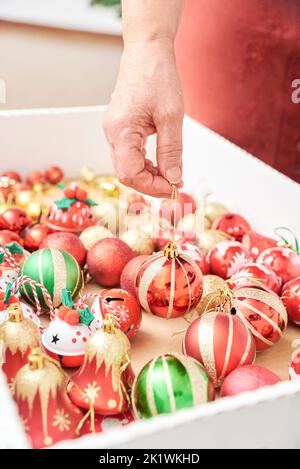 Femmina che afferra una palla da una scatola di ornamenti misti albero di Natale nei classici colori tradizionali di rosso, verde, bianco e oro. Unrecognizab Foto Stock