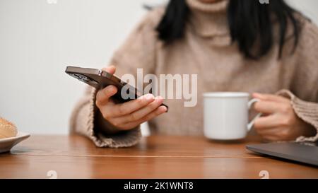 Una donna asiatica che utilizza il suo smartphone per leggere notizie o contenuti online, inviare e-mail, controllare il messaggio mentre sorseggia un caffè mattutino. Immagine ritagliata Foto Stock