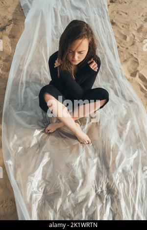 Protezione femminile. Crisi emotiva. Delusione solitudine. Ritratto d'arte di donna sofferente insicura in nero seduto da solo su pellicola di polietilene o Foto Stock