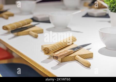 Set di coltelli, tavole e tappetini di sushi di bambù su un tavolo bianco prima della lezione principale di sushi. Concetto di educazione, cucina e intrattenimento. Foto Stock