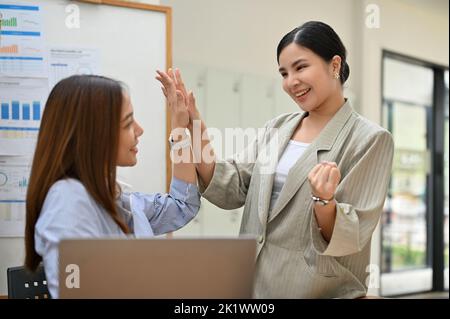 Due donne d'affari asiatiche allegre e allegre, che si danno l'una all'altra in ufficio, celebrando insieme il loro successo. Foto Stock