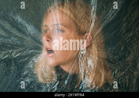 Protezione femminile. Ritratto sfocato. Pressione sociale. Donna bionda ansiosa faccia isolata dietro tessuto rugoso pellicola trasparente in polietilene o Foto Stock