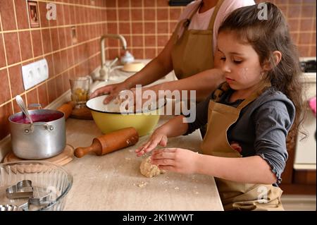 Bella mischievous bambino impastare su un piano di cucina, imparando a cucinare dolci e tartellette con la sua mamma Foto Stock