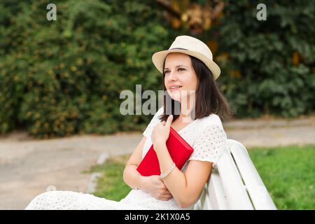 Dreamy Pensive Brunette donna caucasica in abito bianco e cappello di paglia siede sulla panca nel Parco cittadino. La ragazza tiene novella nel libro con copertina rossa. Enj. Femmina Foto Stock