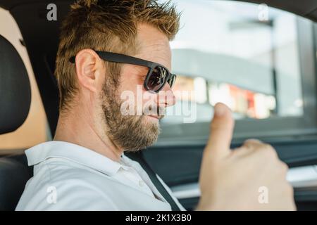 Primo piano ritratto di bel giovane uomo che indossa occhiali da sole in auto mostrando il pollice in su. Foto Stock