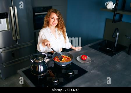 Giovane donna caucasica sorridente cuoce la colazione su una padella. Giovane donna adulta che fa omelette su una stufa a gas. Ragazza che prepara il suo pasto su un Foto Stock