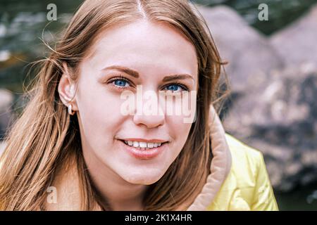 Ritratto di giovane donna attraente con gli occhi blu che guarda la macchina fotografica e sorridente in giacca gialla all'aperto. Foto Stock