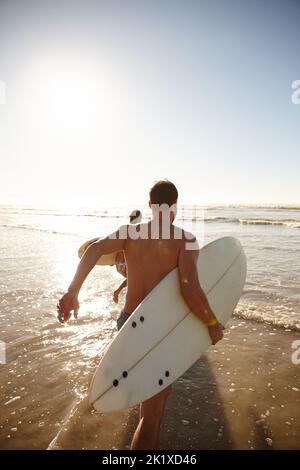 Lascia colpire il surf. Ripresa da dietro di una giovane coppia di surfisti che corrono in mare insieme. Foto Stock