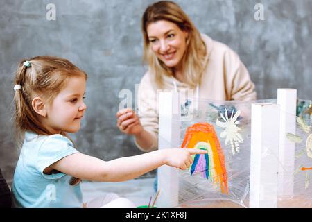 Insegnante femminile con bambina bionda che dipinge arcobaleno su tela oleosa. Divertente apprendimento nel centro di sviluppo per bambini Foto Stock