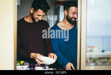 Coppia gay maschio che hanno momento tenero mentre lavano i piatti all'interno della cucina domestica - fuoco sulla faccia sinistra dell'uomo Foto Stock