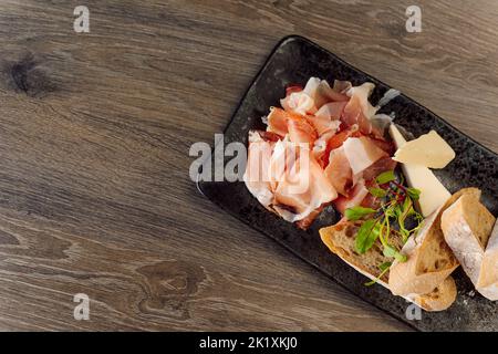 Formaggio, piatto di carne con pane di ciabatta per aperitivo, antipasto su piatto nero su tavolo di legno in caffetteria. Specialità gastronomiche Foto Stock