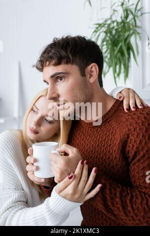 donna bionda che abbraccia il ragazzo in maglione rosso con tazza di tè, immagine stock Foto Stock