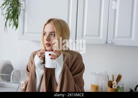 giovane donna bionda in maglione bianco e coperta che tiene una tazza di tè, immagine stock Foto Stock
