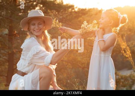 Donna bella e sorridente, madre in cappello e bambina bionda, figlia in abito che si diverte, giocando con le erbe sul prato Foto Stock