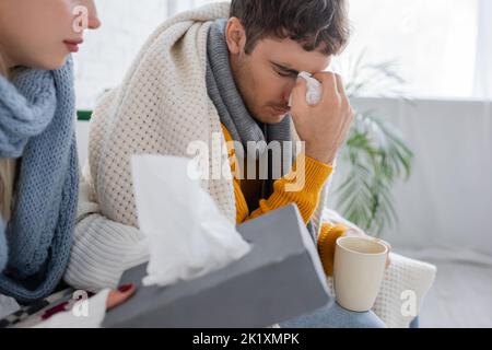 uomo malato con gli occhi chiusi tenendo tovagliolo e tazza vicino alla ragazza con scatola di tessuto, immagine stock Foto Stock