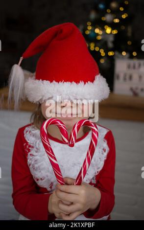 Un bambino sorridente in un berretto rosso di Santa tirato sopra gli occhi tiene le caramelle a forma di cuore nelle sue mani. Il bambino aspetta un miracolo. Natale, Capodanno, Foto Stock