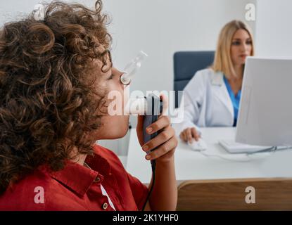 Bambino maschio durante il test di spirometria respiratoria e di funzionalità polmonare utilizzando spirometro medico con il medico che controlla i risultati del test sul monitor Foto Stock