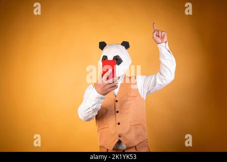 Uomo in maschera di orso panda utilizzando smartphone e puntando il dito su sfondo giallo. Foto Stock