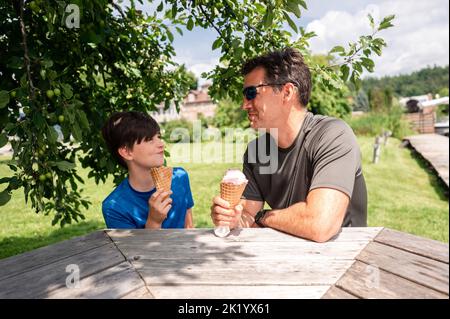 Uomo e ragazzo che mangiano coni gelato al tavolo da picnic il giorno di sole. Foto Stock