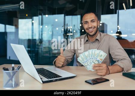 Ritratto di investitore giovane finanziere di successo, uomo sorridente e guardando la macchina fotografica, lavorando all'interno di un moderno edificio per uffici, tenendo in mano denaro in dollari e mostrando pollici positivi in su Foto Stock