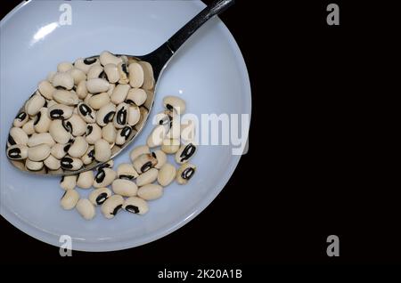 Fagiolo nero essiccato con gli occhi in un cucchiaio da tavola sul piatto, fagioli secchi con i reni su sfondo nero Foto Stock