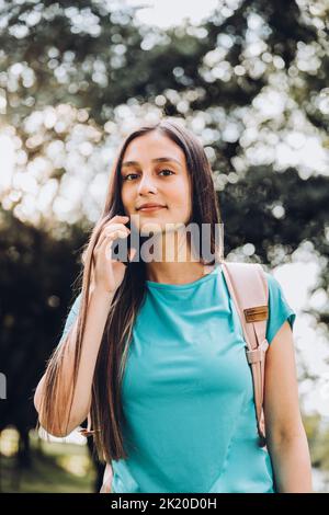 Ragazza studentesca adolescente che indossa una t-shirt acquamarina, facendo una telefonata alla sua famiglia nel parco. Retroilluminazione del sole Foto Stock