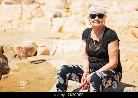 Donna anziana in abiti neri seduti su una roccia vicino al mare Foto Stock