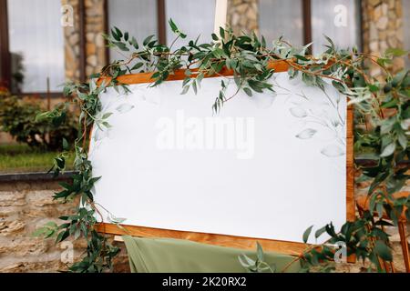 Tavola di legno per matrimoni, mockup, cavalletto per invito, con spazio bianco per un'iscrizione, decorata con verde fresco e stoffa verde. Cornice per matrimoni Foto Stock