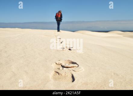 Lasciando il suo segno. Vista posteriore di una giovane escursionista che cammina lungo le dune di sabbia. Foto Stock