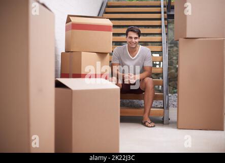 Il cambiamento è una buona cosa. Un giovane felice seduto sulle scale nella sua casa tra scatole di cartone imballate. Foto Stock