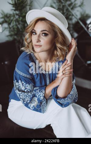 Bella donna bionda elegante in abiti etnici in una camicia ricamata in un cappello bianco all'interno in stile grunge. Nazionale ucraino cl Foto Stock