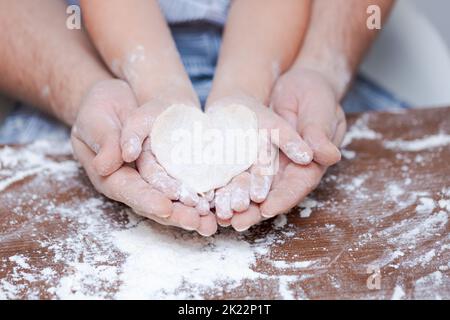 Vista dall'alto la mano del padre e del bambino, tagliare i biscotti a forma di cuore dalla pasta sulle mani sullo sfondo di un tavolo di legno con farina sciolta. Guasto flat. Foto Stock