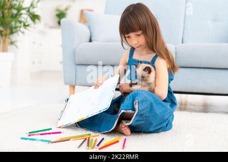 Una ragazza simpatica e divertente in una sundress denim e scopa nei suoi capelli si trova in una stanza su un tappeto e disegna con matite colorate. Accanto a lei c'è un amico a quattro zampe - un piccolo gattino divertente e soffice. Foto di alta qualità Foto Stock