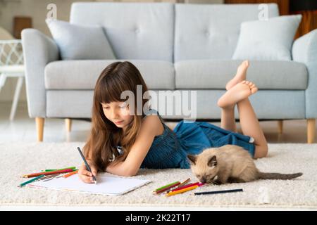 Una ragazza simpatica e divertente in una sundress denim e scopa nei suoi capelli si trova in una stanza su un tappeto e disegna con matite colorate. Accanto a lei c'è un amico a quattro zampe - un piccolo gattino divertente e soffice. Foto di alta qualità Foto Stock