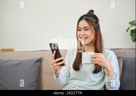 Bella giovane donna asiatica che utilizza il suo smartphone, scorrendo sui suoi social media, leggendo il blog online mentre sorseggia un caffè mattutino e ti rilassa in lei Foto Stock