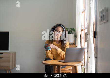 Ritratto di una donna afroamericana che parla in videoconferenza con laptop e cuffie prendendo appunti su blocco note. La brunetta si siede al tavolo dentro Foto Stock