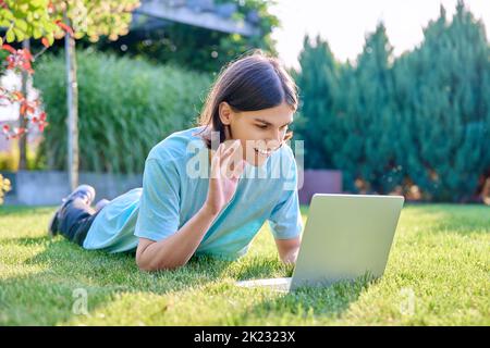 Giovane maschio sdraiato all'aperto sull'erba con un computer portatile che parla con gli amici tramite videochiamata Foto Stock
