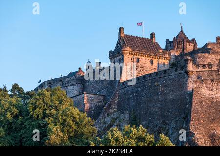 Vista del Castello di Edimburgo che batte una bandiera britannica di Union Jack dalla roccia dell'entroterra con cielo blu chiaro, Scozia, Regno Unito Foto Stock