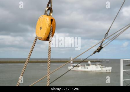 Primo piano di una carrucola e corde su una vecchia nave a vela contro un paesaggio con mare, nuvole e una nave. Foto Stock