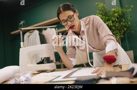 Donna asiatica premurosa che contempla nuove idee di design in uno studio di moda. Sartoriale femminile che lavora su un nuovo modello di abito creativo. Foto Stock
